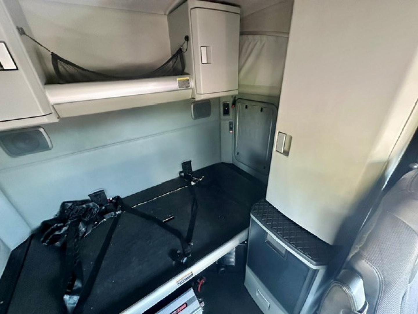 Truck cab interior showing sleeper area with seat belts secured on a flat surface and overhead storage compartments