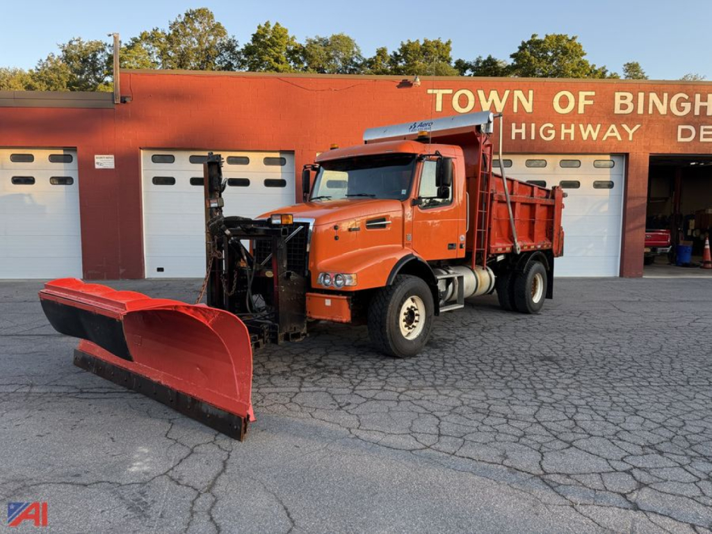 Volvo VHD dump truck with front plow attached, orange cab and dump bed, daytime outdoor lot.