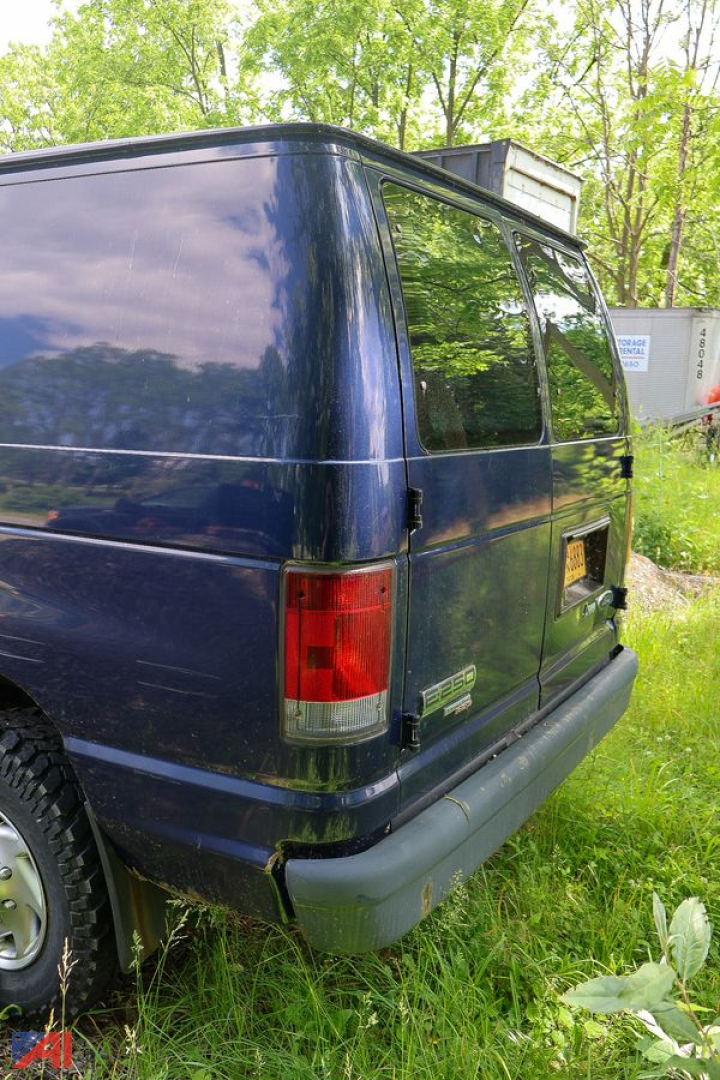 Ford E-250 cargo van rear/side view showing dark blue paint, rear doors, and raised roof; metal bumper; windowed rear passenger side; grassy lot background.