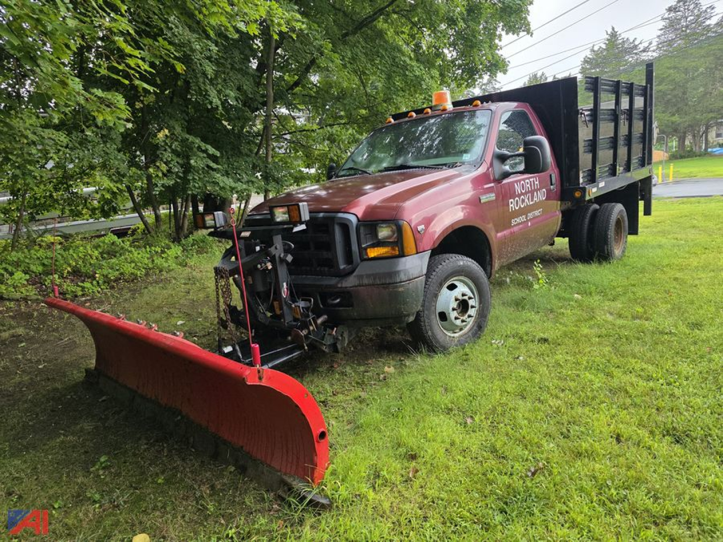 Ford F-350 Super Duty rack truck with a front plow attached, red cab, black dump bed, parked on grass with trees in background.