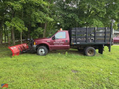 Ford F-350 Super Duty stake truck with a front plow and a flatbed/utility bed labeled North Rockland School District, side view showing plow attachment and black fenced utility bed.