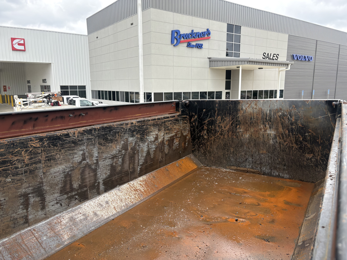Mack GR64F dump truck bed interior showing rust-stained steel walls and floor, dealership building with Brackner? signage in background.