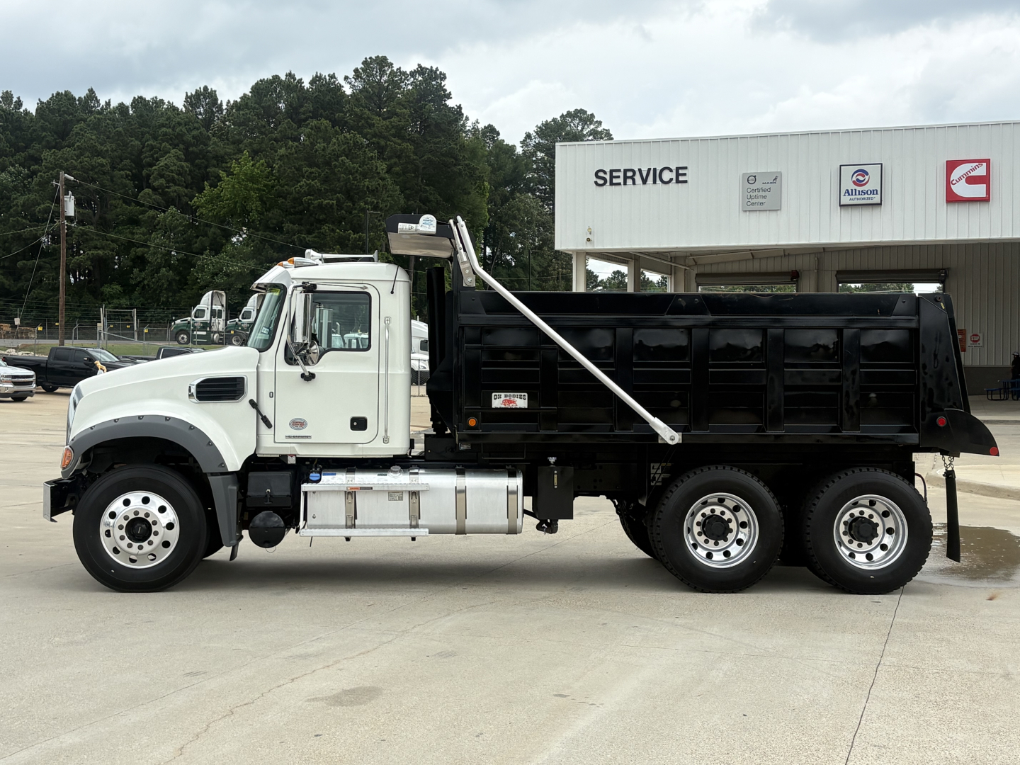 Dump truck side view showing Mack GR64F with white cab and black dump bed, service building in background.