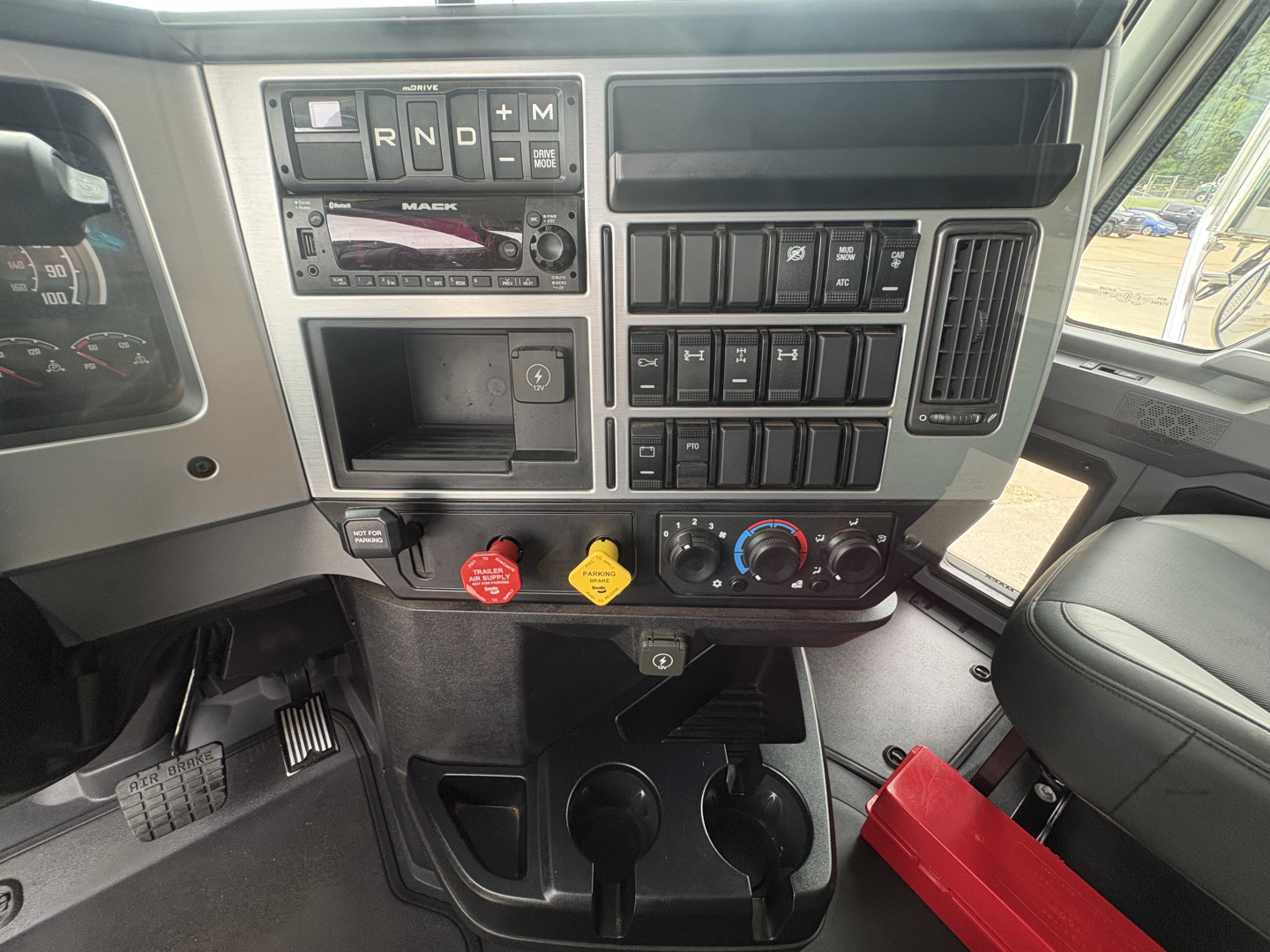 Dashboard of a Mack truck cab showing transmission gear selector, control switches, and knobs with labeled buttons and two cup holders.