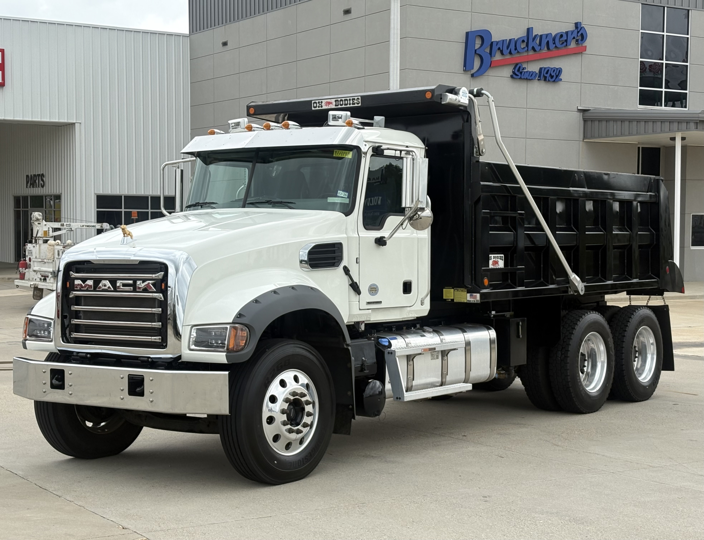 Mack GR64F dump truck in white cab with black dump bed, front grille MARCO MACK, parked in lot in front of dealership building.