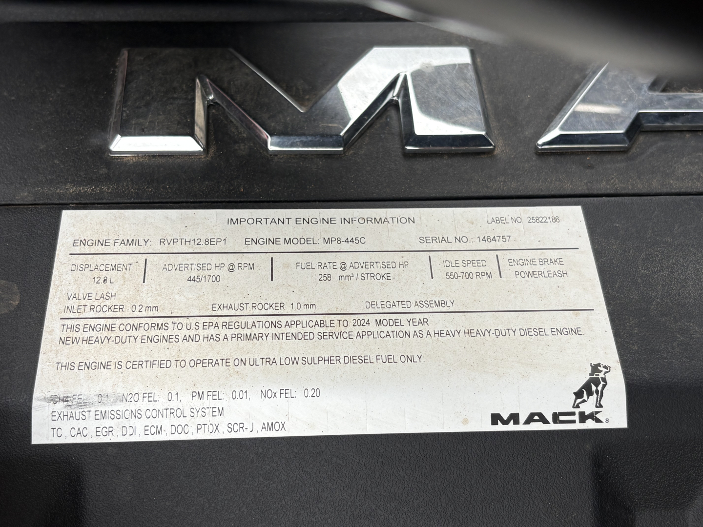 Mack engine badge and label with important engine information plate showing engine family, model, serial number, and powertrain specs on a Mack GR64F truck engine cover.