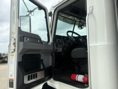 Interior of a 2025 Mack GR64F dump truck cab showing steering wheel, dashboard, and door with small storage compartment. Red fire extinguisher mounted near the floor and open door frame.