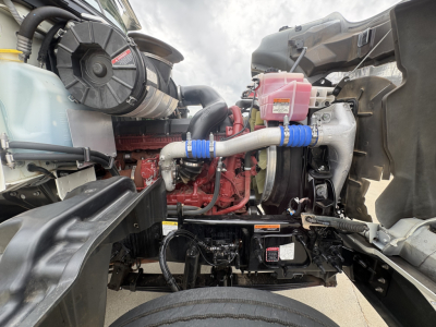 Mack GR64F engine bay showing coolant reservoir, pink anti-freeze bottle, red engine block, hoses with blue clamps, air intake, and hydraulic components.