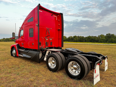 Kenworth T680 conventional sleeper truck in red, rear three-quarter view showing dual rear axles and pickup of mud flaps with MHC logo.