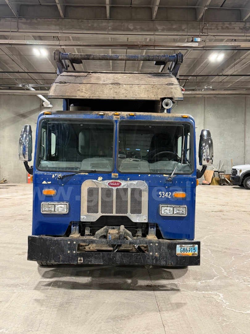 Front view of a blue 2017 Peterbilt 320 garbage truck showing grill, headlights, bumper, and raised compactor in an indoor facility.