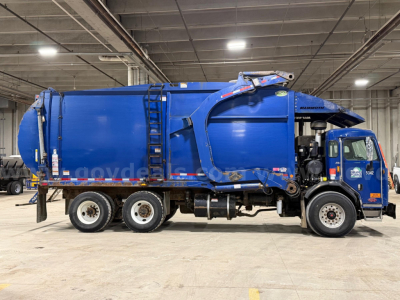 Peterbilt 320 garbage truck in a warehouse, side view showing blue body, rear compactor, and dual rear axles.