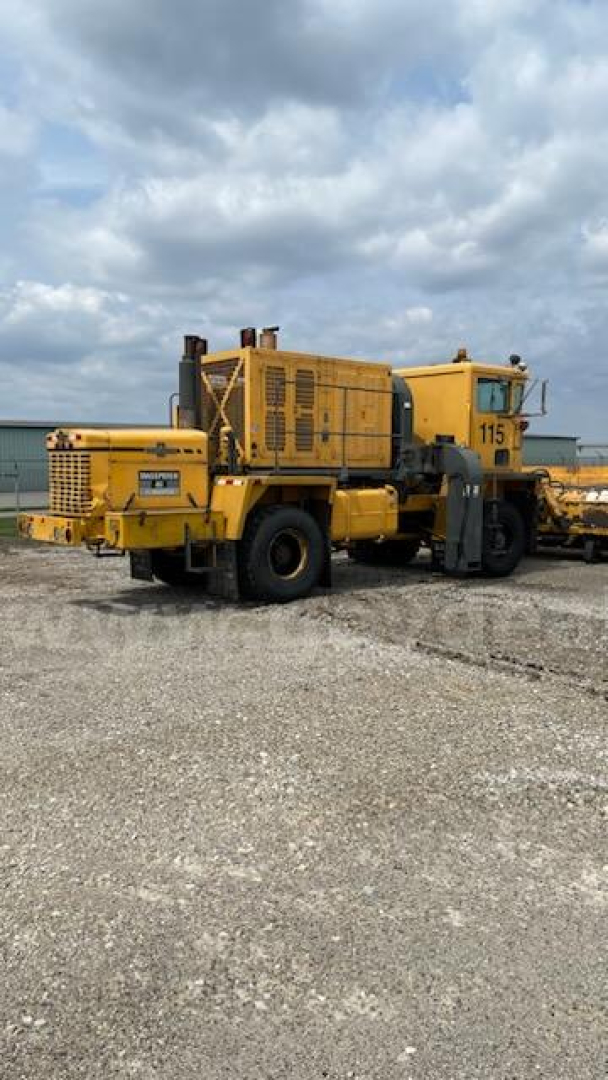 Yellow Oshkosh heavy equipment grader/truck with large rear wheels and exhaust stack in a gravel lot, model number 115 visible on side.