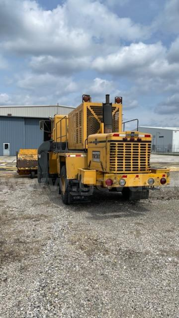Front-left view of a yellow Oshkosh Broom road recycler with large rear engine compartment and tracks visible, showing heavy-duty machinery and rear grilles.