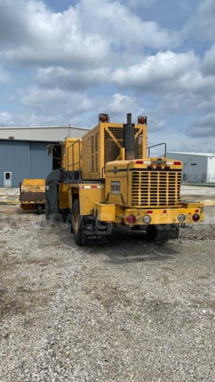 Front-left view of a yellow Oshkosh Broom road recycler with large rear engine compartment and tracks visible, showing heavy-duty machinery and rear grilles.
