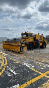 Back view of a yellow Oshkosh broom street sweeper on a gravel lot with large front roller and cabin labeled '115'