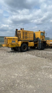 Yellow Oshkosh heavy equipment grader/truck with large rear wheels and exhaust stack in a gravel lot, model number 115 visible on side.