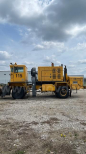 1990 Oshkosh Broom with yellow industrial machine, side view showing numbered cab 115 and engine unit, on a gravel lot.