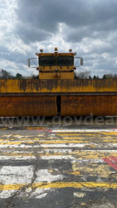 Front view of a yellow Oshkosh snow plow or road grader with large rusty roller blade and cloudy sky.
