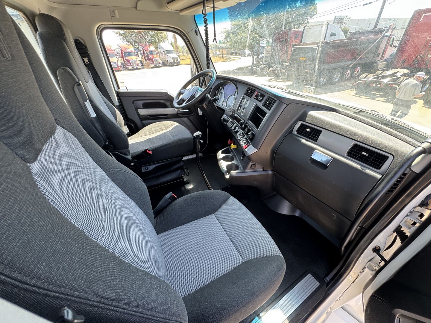 Kenworth T680 cabin interior view showing steering wheel, dashboard with gauges, driver seat and passenger seat, Beige/gray upholstery, center console, and entry door.