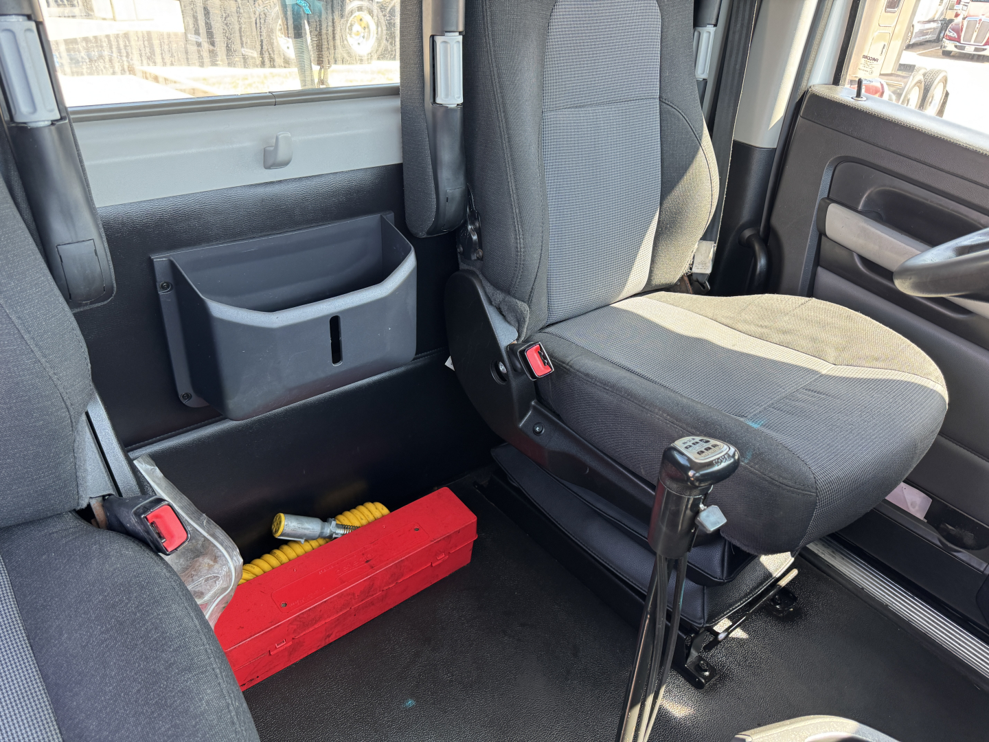Cab interior of a Kenworth T680, captain seat with gear shifter to the right, red tool tray on floor, plastic storage pocket on side wall, gray upholstery, badge area not visible.