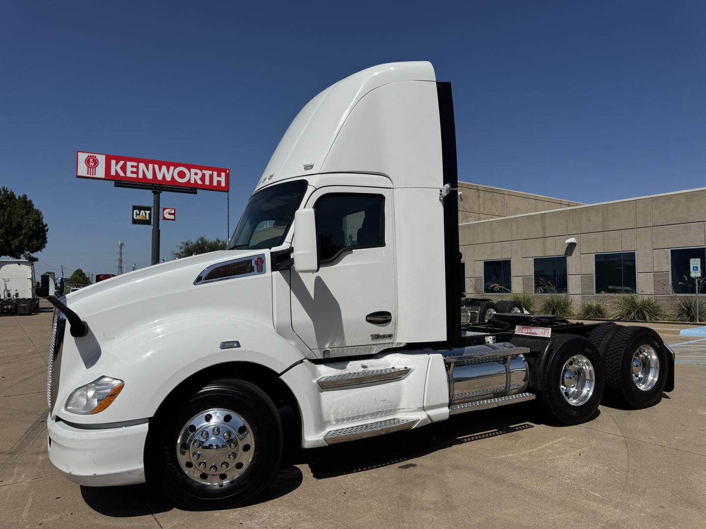 Kenworth T680 conventional daycab tractor showing side view with white paint, chrome fuel tanks, dual rear axles, and Kenworth sign in background.
