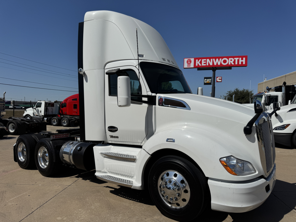 Kenworth T680 tractor in white, side view showing cab, front wheel, and dual rear axles; Kenworth dealership signage in background.