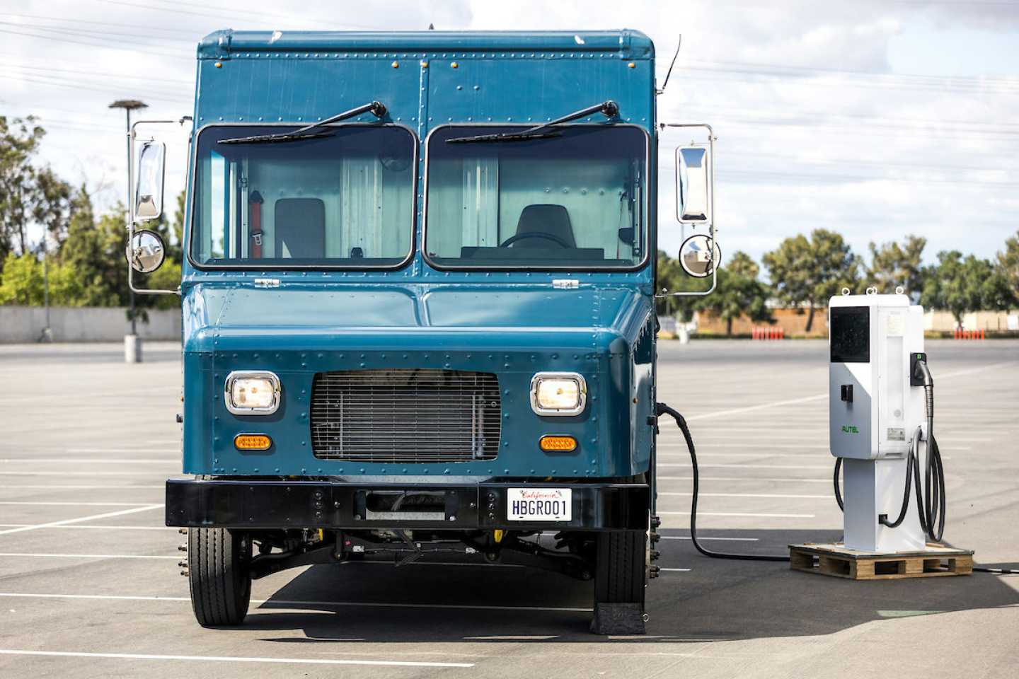 Harbinger S656 step van front view in blue with charging station on right, front grille, headlights, and windshield wipers visible.