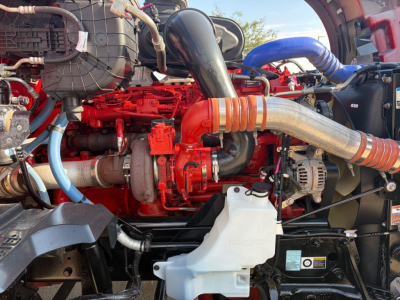 Kenworth T680 engine bay showing red diesel engine, large intake tube, hoses, alternator, coolant reservoir, and various metal components.