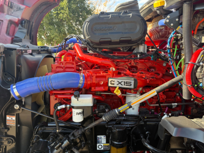 Kenworth T680 engine bay showing red engine with X15 label, blue hose, white reservoir, and various hoses and wiring.