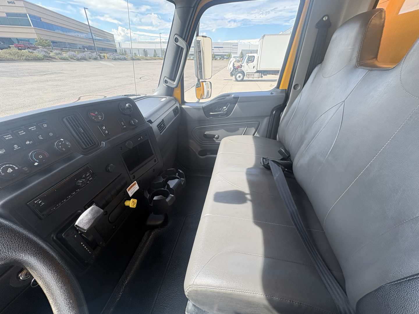 Interior cab of a 2021 International MV607 truck showing dashboard controls, steering wheel, and grey bench seat with seat belt. Exterior view through window shows yard and buildings.