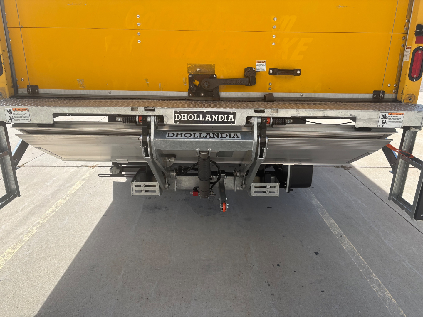 Vehicle rear showing Dhollandia tow hitch and lift gate on a yellow box truck with visible bumper components.
