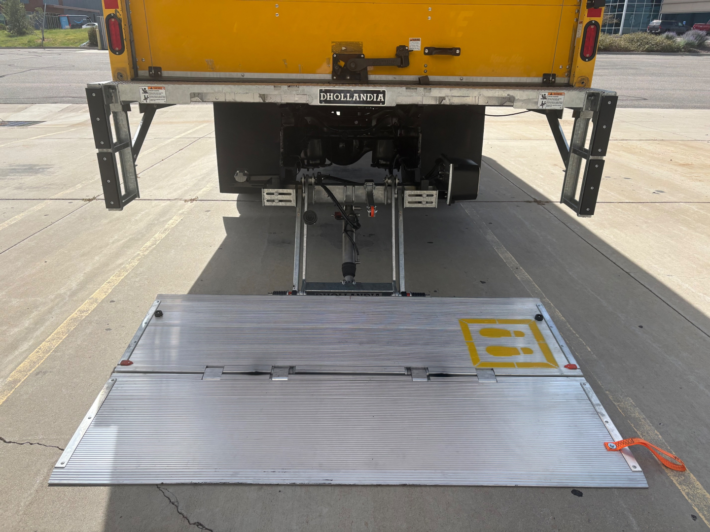 Truck rear with liftgate extended, ISO view of tail area and platform showing 'Dhollandia' label, yellow cargo box, chassis and mechanical assemblies visible.