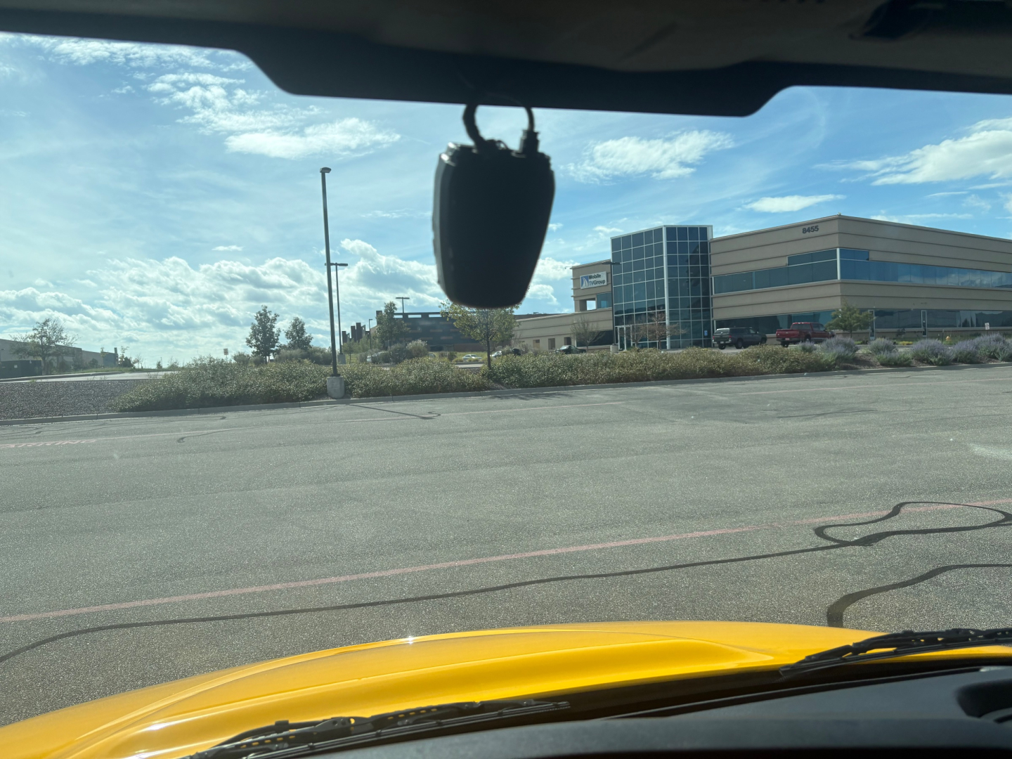 Side view of a yellow box truck hood from the inside of the cab, with a gray hanging air freshener or device, and a modern building in the background.