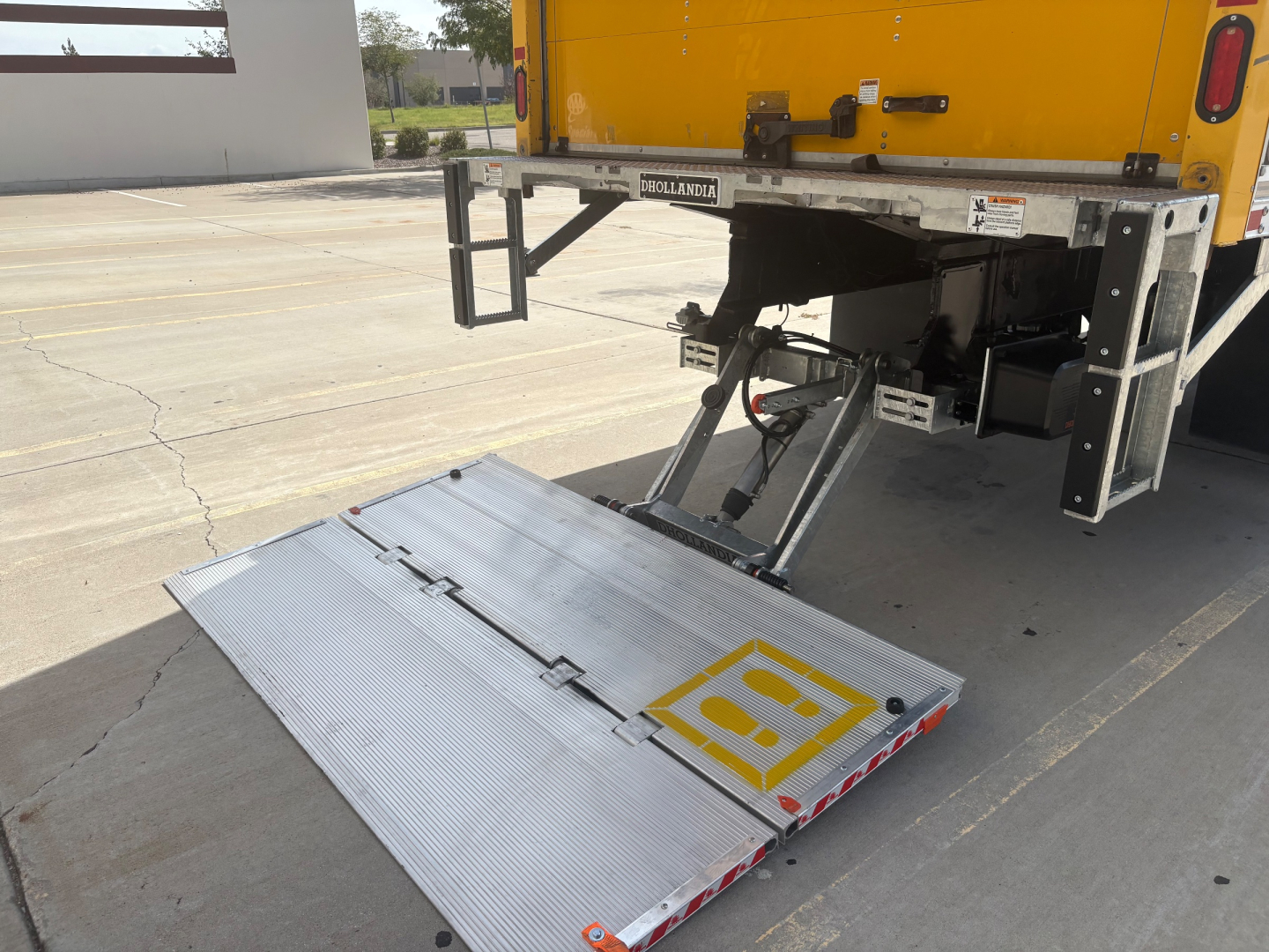 Truck rear with liftgate extended, yellow box truck body, silver aluminum fold-down liftgate platform, hydraulic arms visible, some warning decals on platform