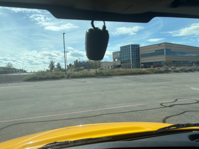 Vehicle interior view of a truck from the windshield; a black hanging object in the upper center; exterior shows a yellow hood and a parking lot with a modern building in the background.
