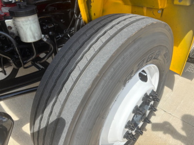Front wheel of a 2021 International MV607 truck showing tire and wheel with hub bolts; yellow chassis in background; visible engine bay components and reservoir.