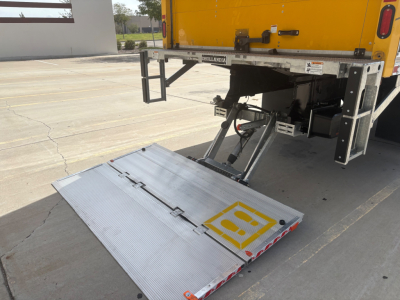 Back of a box truck with a lowered aluminum liftgate. Yellow truck body visible; hydraulic arms and platform; metal safety rails; nozzles and wiring visible around the rear.