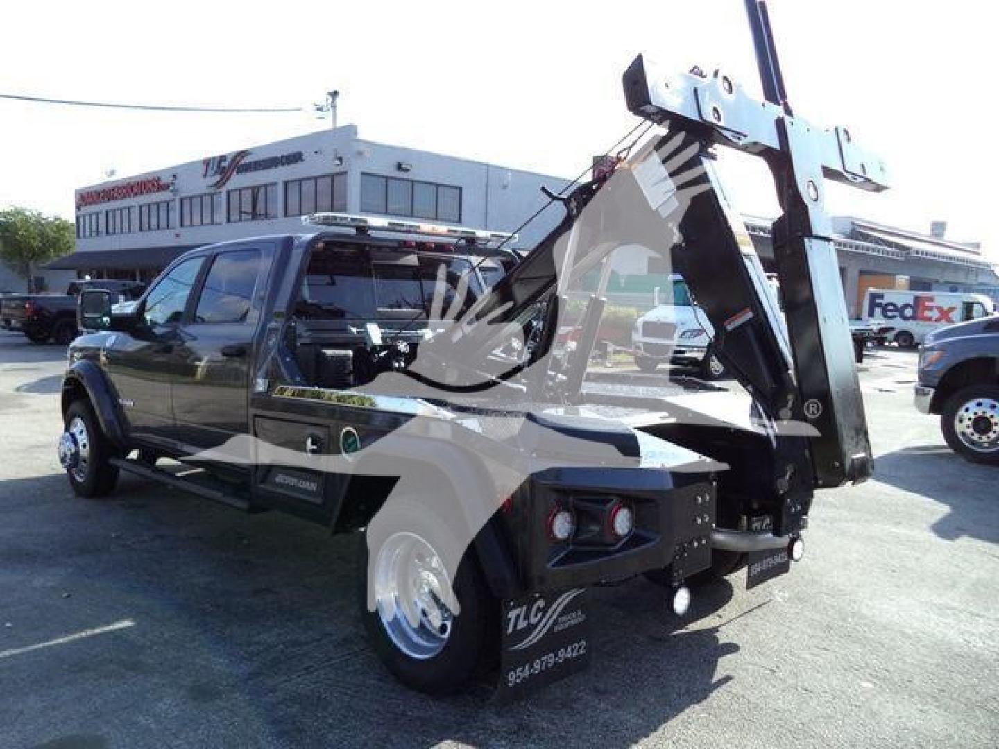 Ram 5500 tow truck side view with crane/winch system, showing operator cab and heavy-duty rear equipment. Visible text includes 'J25602' and 'FedEx' in background, plus dealership signage on building.