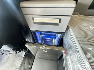 Truck interior storage area with open center console drawer and blue-lit organizer cabinet in a Mack Anthem, showing a metal drop-down drawer under a gray upper console.