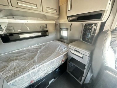 Truck cab interior showing sleeper berth with plastic-wrapped mattress, overhead storage cabinets, control panel, window, and side console.