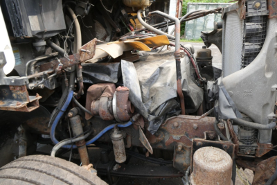 Engine bay showing damaged front end components of a Freightliner CL112 with exposed engine wiring, collapsed hoses, and crumpled metal fender area.