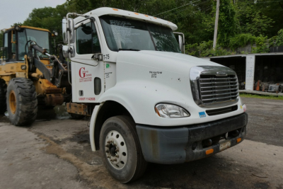 White 2006 Freightliner CL112 conventional daycab truck showing front grille, headlights, bumper, and side decals including stock number and VIN placards