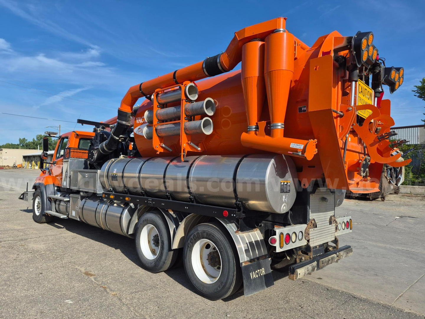 Side view of a Freightliner 114SD vacuum truck with large orange pumping unit mounted on bed and multiple cylindrical tanks; visible rear wheels and metal steps.