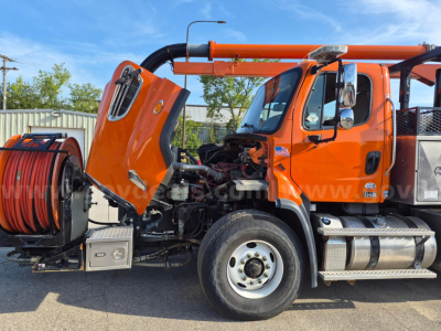 Orange Freightliner 114SD vacuum truck with hood open, showing engine and reels; side mirrors, chrome fuel tank, and step; front wheel and bumper visible.