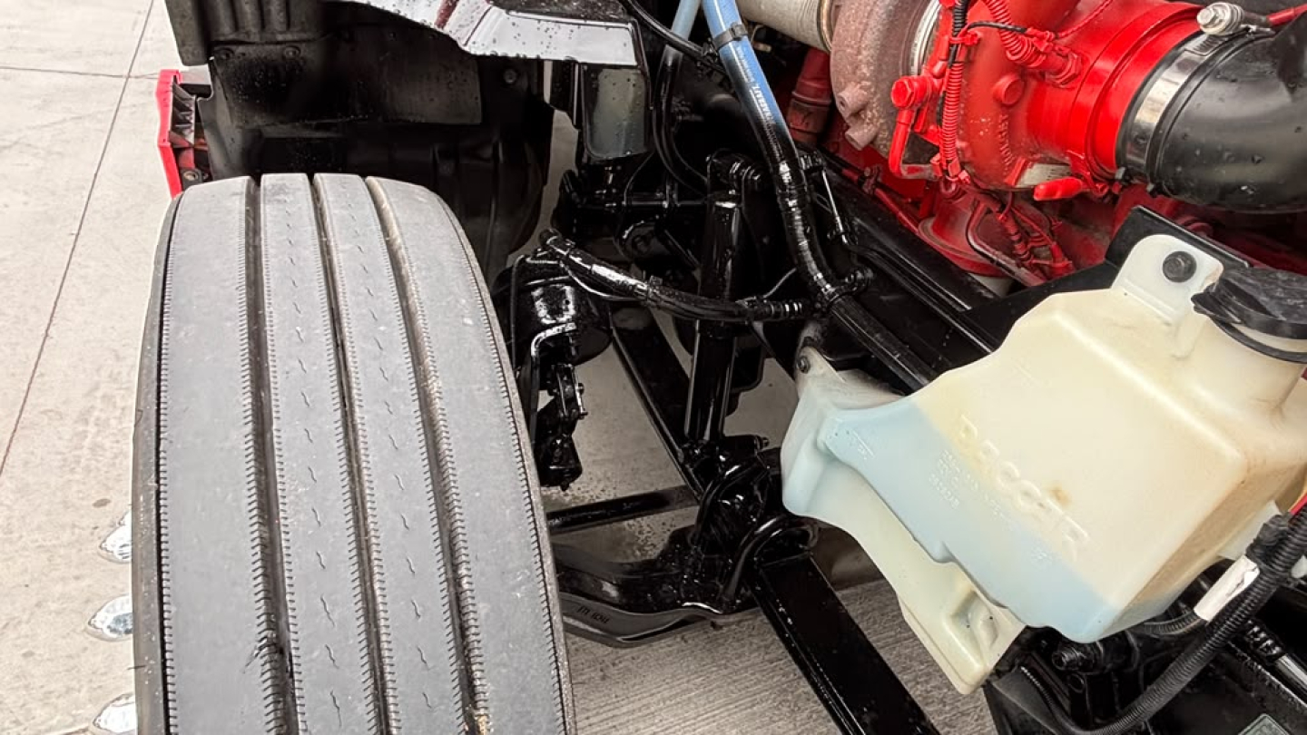 Truck front wheel well and engine bay components: visible coolant reservoir with cap, red engine block, black chassis, and hoses. No legible model text visible on image.