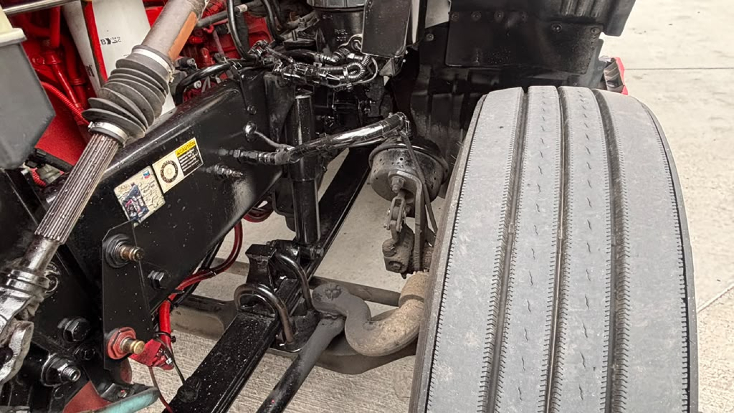 Kenworth T680 engine bay and front suspension showing exhaust manifold and large front tire from a used conventional sleeper truck.