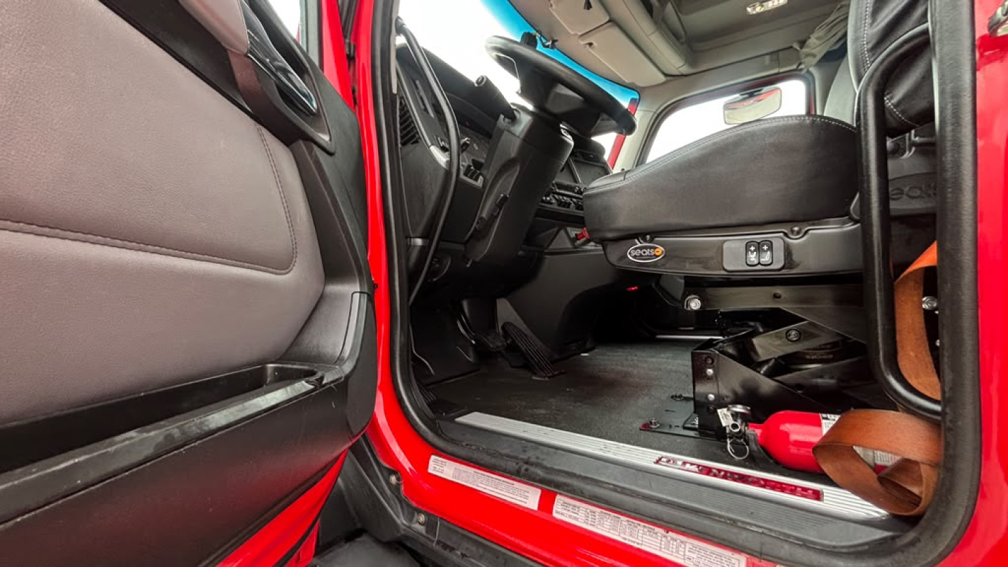 Cab interior of a 2023 Kenworth T680 in-view showing driver seat, steering wheel, pedals, and door sill with VIN/stock stickers nearby.