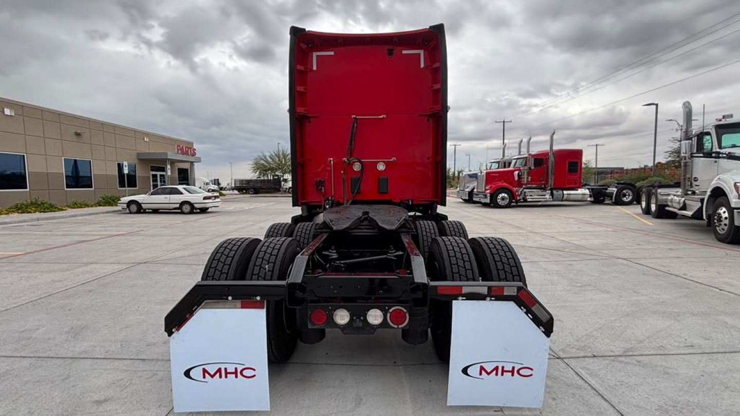 Truck rear view showing fifth wheel and dual rear axles of a Kenworth T680 sleeper, red cab back, white mud flaps with MHC logos, parking lot background.