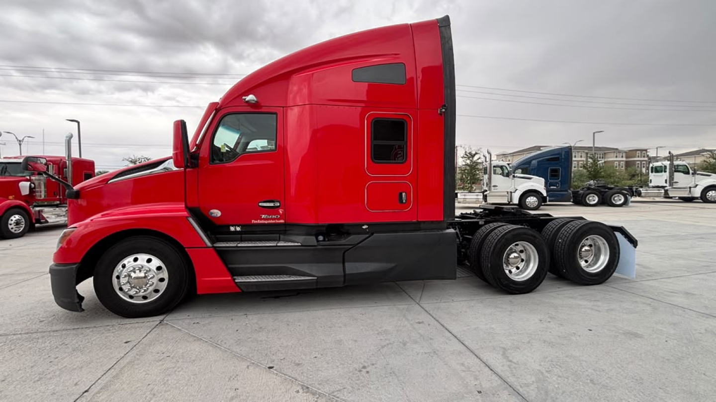 Kenworth T680 semi-truck in red with sleeper cab, side view showing front grille, hood, and triple rear axles on a concrete lot.