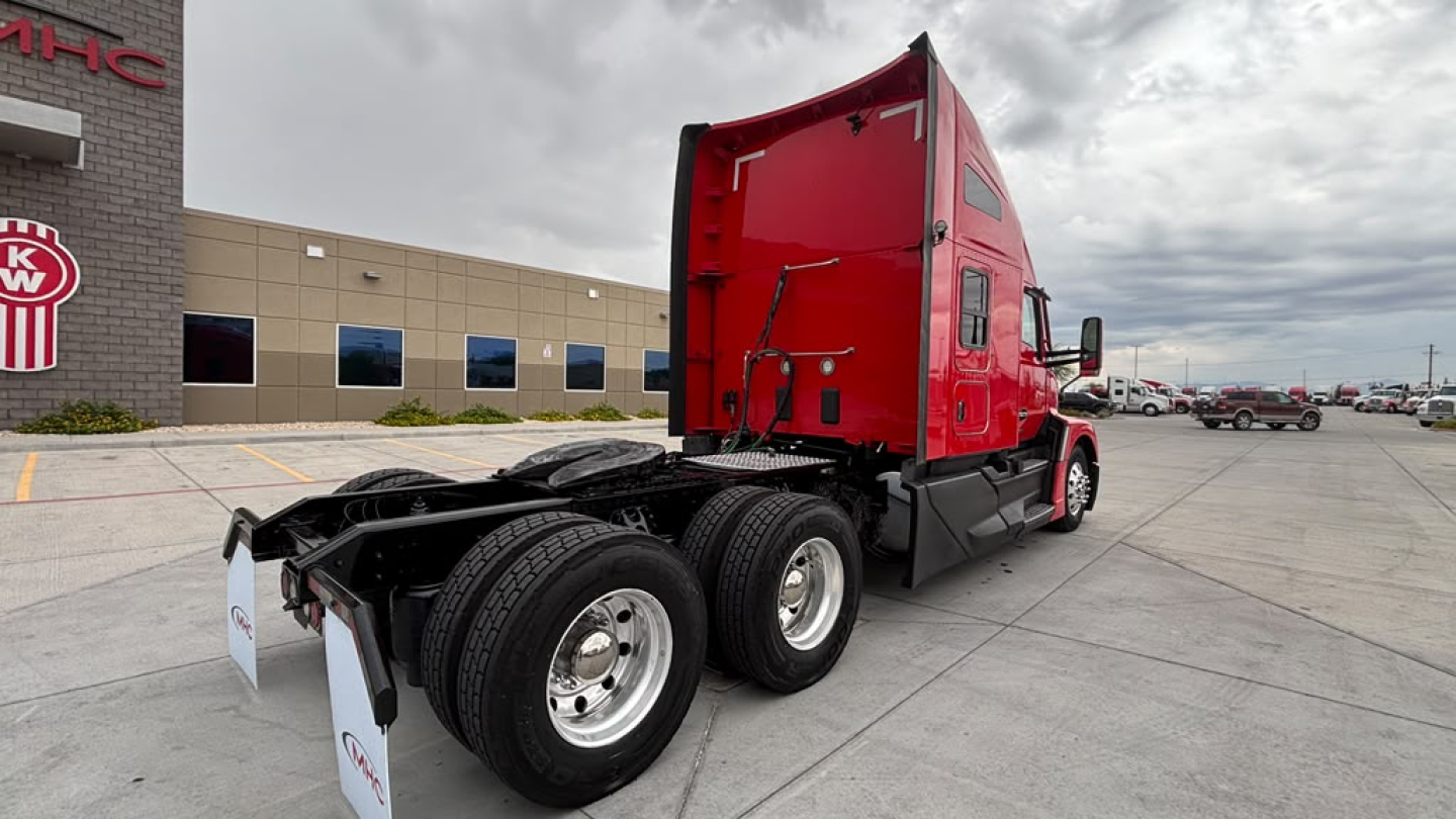 Kenworth T680 tractor trailer cab in red with exposed rear chassis and dual rear axles, parked in lot near dealership.